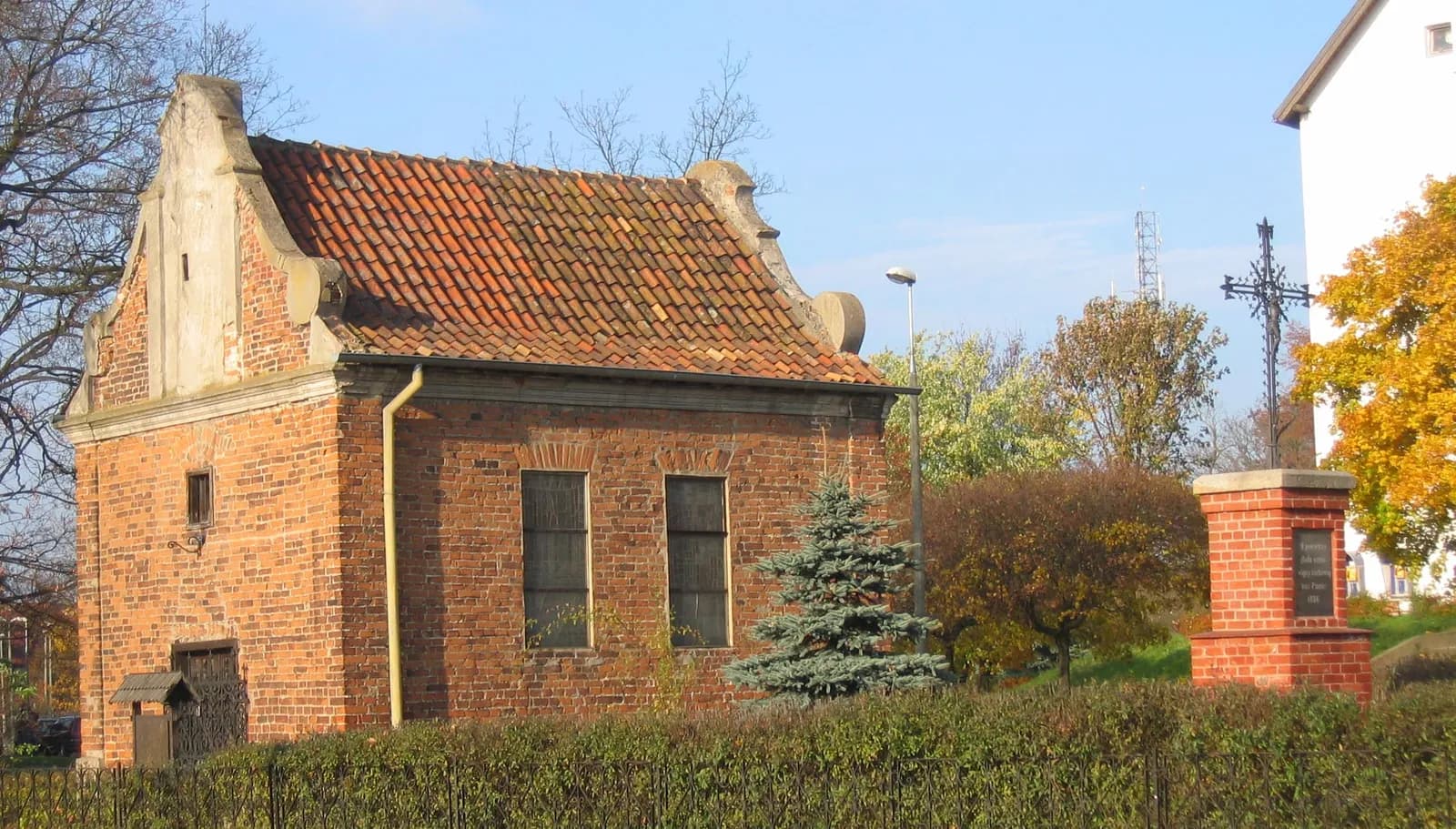 A historic brick building with a tiled roof stands behind a hedge, with a small fir tree and a brick monument topped with a cross nearby.