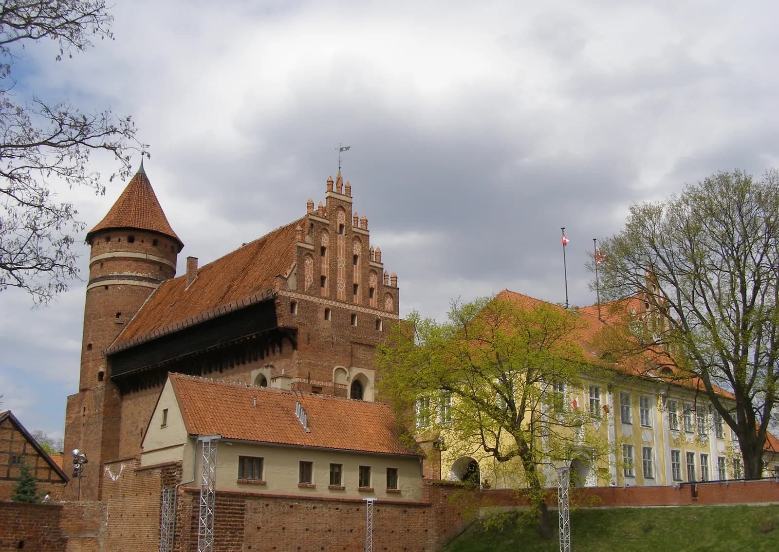 A large red brick castle with a conical roof tower on the left and a large gabled section to the right, next to a lighter colored building with a red roof, all under a cloudy sky.